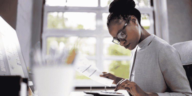 Mulher sentada, em frente à uma mesa, utilizando uma calculadora e um computador, enquanto segura documentos, fazendo cálculos.