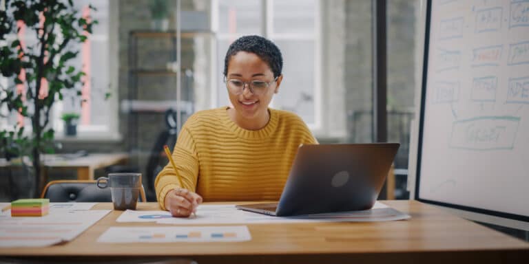 Empreendedora está organizando o seu planejamento para abrir uma empresa, em seu escritório, que conta com mesa, quadro de anotações e uma planta ao fundo.