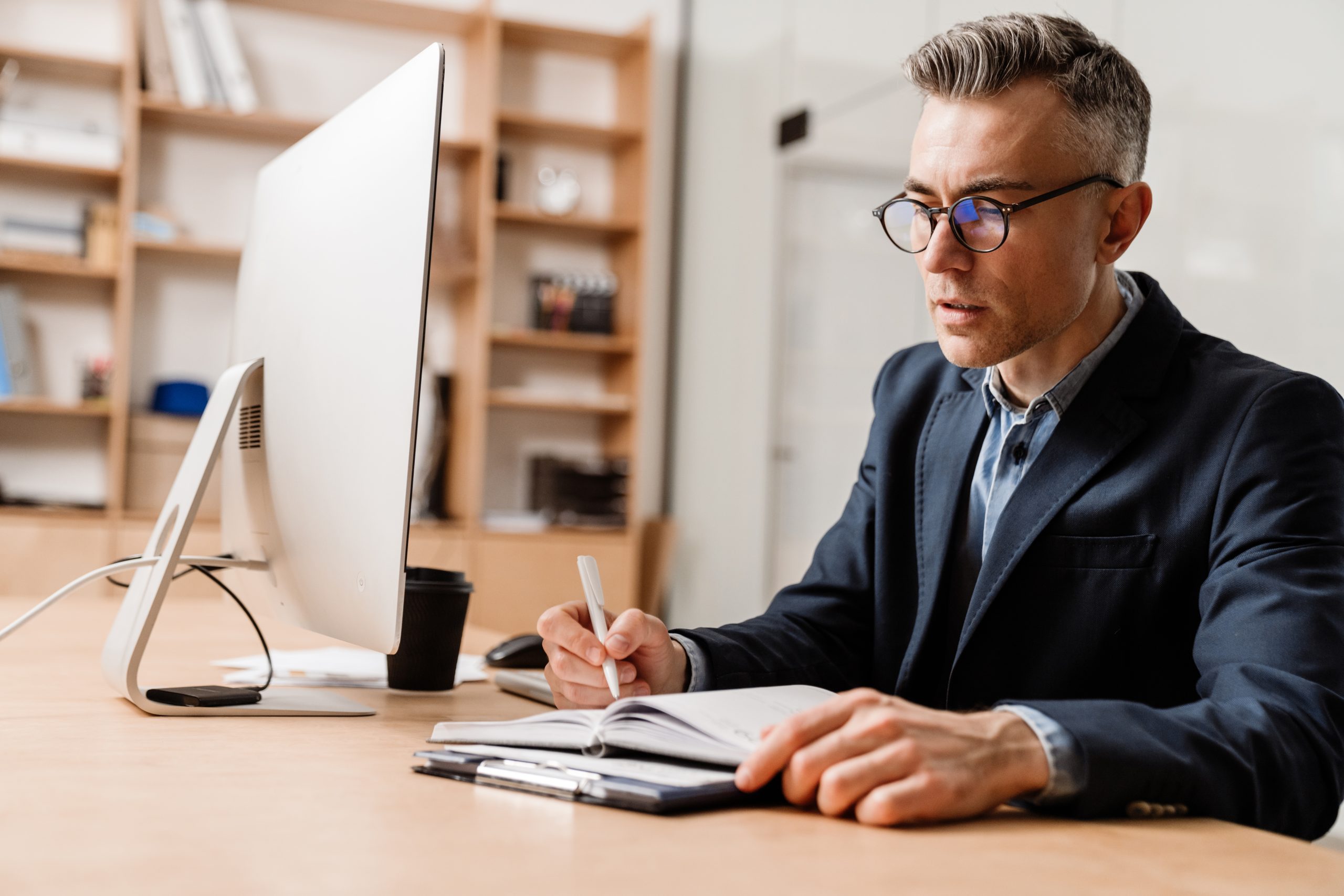 Serious grey man working with computer and planner while sitting at table in office