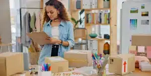 Mulher faz anotações numa prancheta; e em uma mesa há diversas caixas de papelão, em imagem que ilustra artigo sobre as ferramentas de cobrança da conta PJ da Cora.