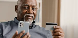 Homem segura o celular com uma mão e o cartão de crédito com a outra, para conferir as informações, em imagem que ilustra artigo sobre o cartão de crédito PJ da Cora.
