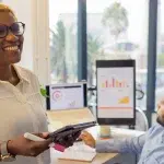 Mulher sorri enquando segura um tablet e uma caneta. Ao fundo, homem trabalha em um computador, com gráficos na tela, em imagem que ilustra artigo sobre como a automatização para fazer cobranças em massa melhora gestão financeira de empresas.