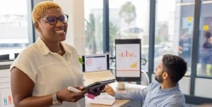 Mulher sorri enquando segura um tablet e uma caneta. Ao fundo, homem trabalha em um computador, com gráficos na tela, em imagem que ilustra artigo sobre como a automatização para fazer cobranças em massa melhora gestão financeira de empresas.
