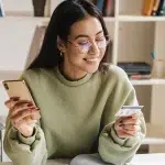Mulher de óculos sorri ao ler informações em um cartão, enquanto segura o celular com a outra mão, em imagem que ilustra artigo sobre contas PJ sem tarifa.