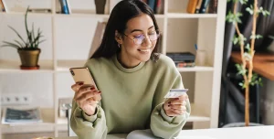 Mulher de óculos sorri ao ler informações em um cartão, enquanto segura o celular com a outra mão, em imagem que ilustra artigo sobre contas PJ sem tarifa.