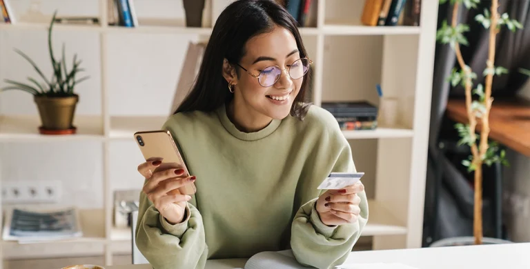 Mulher de óculos sorri ao ler informações em um cartão, enquanto segura o celular com a outra mão, em imagem que ilustra artigo sobre contas PJ sem tarifa.
