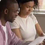 Homem e mulher analizam papelada e utilizam um computador em imagem que ilustra este artigo sobre os documentos necessários para abrir uma conta PJ.