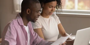 Homem e mulher analizam papelada e utilizam um computador em imagem que ilustra este artigo sobre os documentos necessários para abrir uma conta PJ.