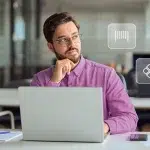 Homem pensa em quanto trabalha em seu laptop. Ao seu redor há ícones financeiros, em fotomontagem que ilustra este artigo sobre boletos Pix como forma de cobrança para empresas.