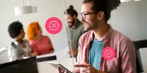 Fotomontagem de um homem utilizando um tablet para ilustrar artigo sobre juros flexíveis. Ao seu redor há ícones de porcentagem e, ao fundo, três pessoas conversam.