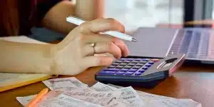 Mão de uma pessoa segurando uma caneta e utilizando uma calculadora. Na mesa, ao redor, diversos recibos e um laptop, em imagem que ilustra guia completo do IRPJ.