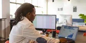 Homem observa gráficos em um laptop. Próximo a ele também tem uma outra tela, com diversos dados em uma planilha, em imagem que ilustra artigo sobre qual é a melhor plataforma de cobranças para empresas.