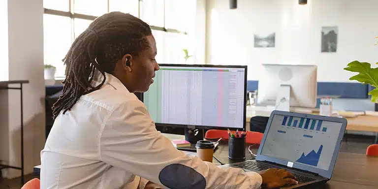 Homem observa gráficos em um laptop. Próximo a ele também tem uma outra tela, com diversos dados em uma planilha, em imagem que ilustra artigo sobre qual é a melhor plataforma de cobranças para empresas.