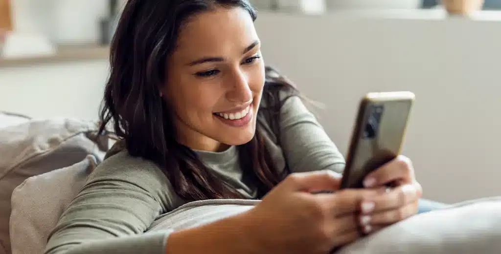 Mulher sorri ao digitar em seu smartphone, em imagem que ilustra artigo sobre com abrir uma conta PJ.