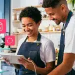 Duas pessoas sorrim enquanto observam um tablet em uma panificadora. Na fotomontagem, que ilustra artigo sobre boletos recorrentes e Pix Automático, há também ícones financeiros.