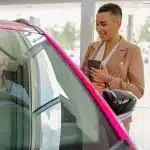 Homem sentado no carro dentro da concessionária enquanto a vendedora sorri do lado de fora, em imagem que ilustra artigo sobre desconto PJ para comprar veículos.