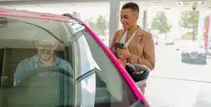 Homem sentado no carro dentro da concessionária enquanto a vendedora sorri do lado de fora, em imagem que ilustra artigo sobre desconto PJ para comprar veículos.