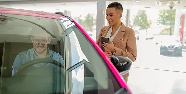 Homem sentado no carro dentro da concessionária enquanto a vendedora sorri do lado de fora, em imagem que ilustra artigo sobre desconto PJ para comprar veículos.