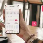 Fotomontagem da tela do app da conta PJ da Cora em um smartphone segurado por uma pessoa, com diversos ícones que representam diferentes usuários.