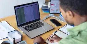 Homem faz anotações em um caderno enquanto observa uma planilha na tela de seu laptop, em imagem que ilustra artigo sobre conciliação bancária automática.