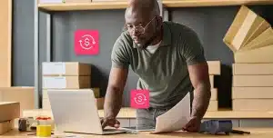 Homem, em pé, utiliza laptop que está sobre uma mesa enquanto segura documentos com a outra mão. Ao fundo, há caixas de papelão, e ícones financeiros completam a fotomontagem que ilustra artigo sobre como fazer simulações reais de crédito para pequenas empresas.