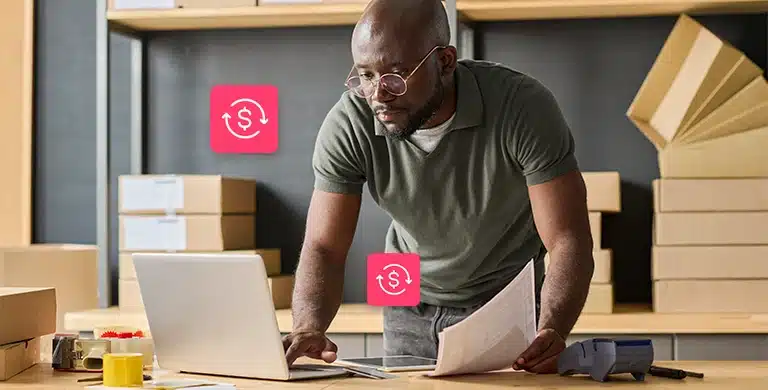 Homem, em pé, utiliza laptop que está sobre uma mesa enquanto segura documentos com a outra mão. Ao fundo, há caixas de papelão, e ícones financeiros completam a fotomontagem que ilustra artigo sobre como fazer simulações reais de crédito para pequenas empresas.