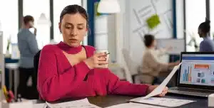 Mulher sentada à mesa em um escritório, segurando uma xícara e um documento impresso, com notebook aberto exibindo gráficos ao lado.