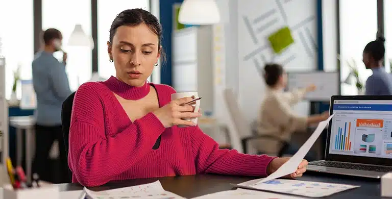 Mulher sentada à mesa em um escritório, segurando uma xícara e um documento impresso, com notebook aberto exibindo gráficos ao lado.