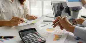 Equipe analisando relatórios financeiros impressos, gráficos de desempenho e planilhas sobre a mesa, com calculadora e notebook, em atividade de planejamento financeiro.