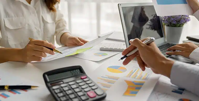 Equipe analisando relatórios financeiros impressos, gráficos de desempenho e planilhas sobre a mesa, com calculadora e notebook, em atividade de planejamento financeiro.