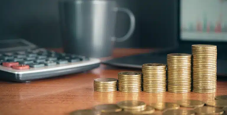 Pilha de moedas organizadas em ordem crescente sobre uma mesa, com calculadora, xícara e notebook desfocados ao fundo.