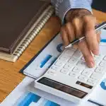 Mão utilizando uma calculadora sobre relatórios com gráficos, com caderno e caneta sobre a mesa.