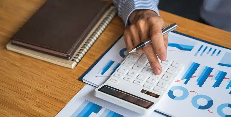 Mão utilizando uma calculadora sobre relatórios com gráficos, com caderno e caneta sobre a mesa.