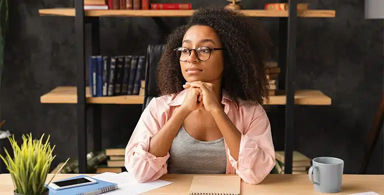 Mulher parece pensativa, com os cotovelos apoiados em uma mesa e o queixo apoiado nas mãos, em imagem que ilustra artigo sobre o que avaliar antes de abrir uma conta PJ para prestador de serviço.