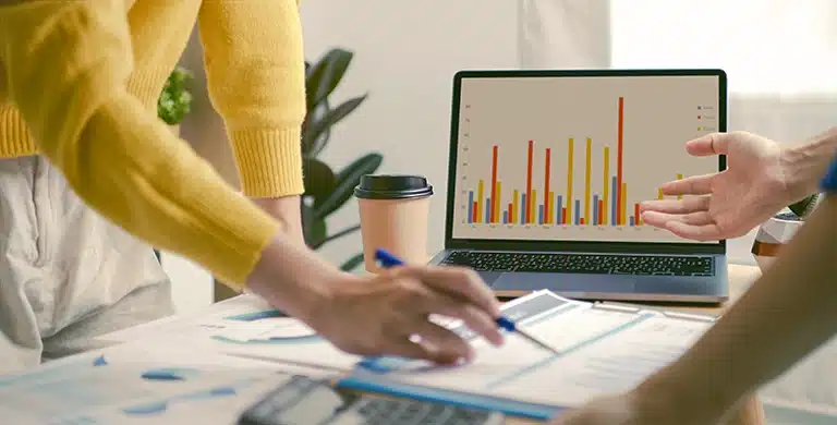 Mãos de duas pessoas diferentes sobre uma mesa com documentos impressos; uma segura uma caneta sobre os papéis e a outra faz um gesto em direção a um notebook ao fundo, que exibe gráficos de barras.