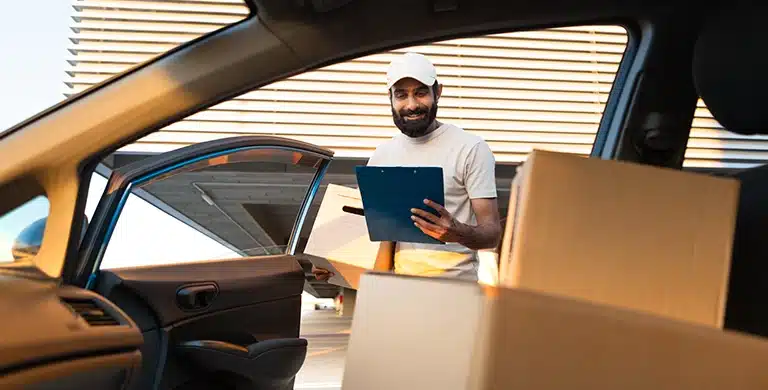 Pessoa em pé ao lado de um carro aberto, segurando uma prancheta, com caixas de papelão no interior do veículo.
