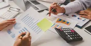 Imagem que ilustra artigo sobre a Reforma Tributária 2026, com pessoas analisando gráficos e relatórios financeiros em uma mesa de trabalho, ao lado de calculadora, notebook e documentos impressos.