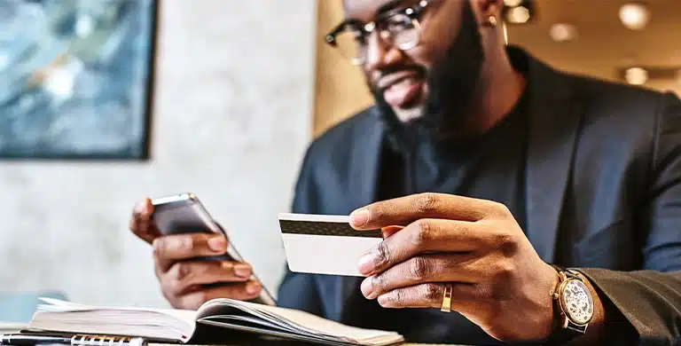 Pessoa segurando um cartão e um smartphone, sentada à mesa, com um caderno aberto à frente