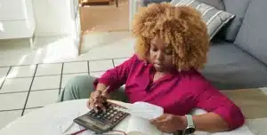 Mulher sentada no chão, apoiada em um sofá, com calculadora e papéis sobre uma mesinha, em imagem que ilustra artigo sobre se receita de pessoa física conta para limite do MEI em 2026.