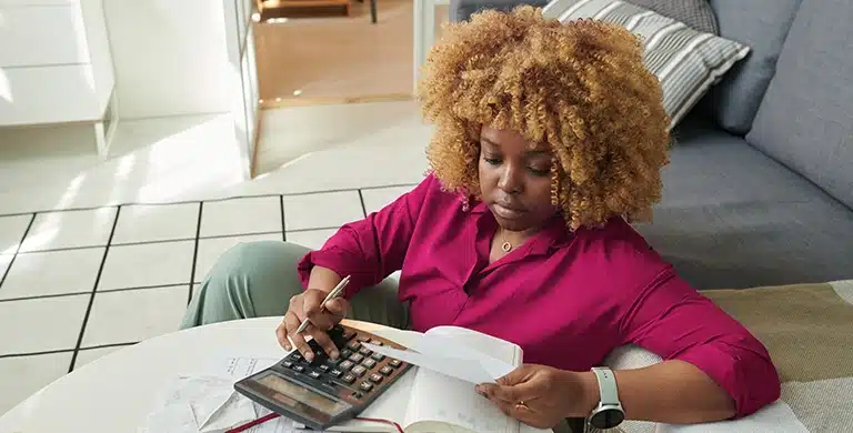 Mulher sentada no chão, apoiada em um sofá, com calculadora e papéis sobre uma mesinha, em imagem que ilustra artigo sobre se receita de pessoa física conta para limite do MEI em 2026.