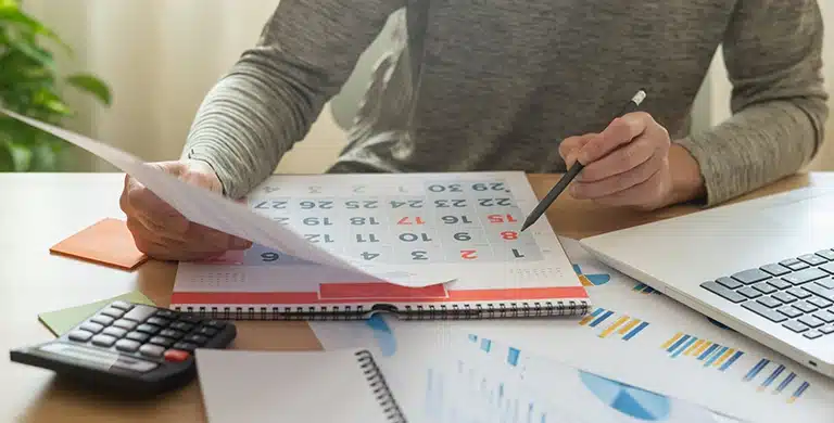 Pessoa com as mãos sobre a mesa, com papel, caneta, calendário, calculadora, laptop e gráficos, em imagem que ilustra artigo sobre Calendário de obrigações do PJ para 2026.