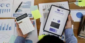 Pessoa segurando tablet com gráficos financeiros e calculadora sobre mesa com relatórios impressos, em imagem que ilustra artigo sobre automatizar pagamentos recorrentes na conta PJ.