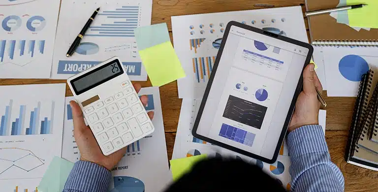 Pessoa segurando tablet com gráficos financeiros e calculadora sobre mesa com relatórios impressos, em imagem que ilustra artigo sobre automatizar pagamentos recorrentes na conta PJ.