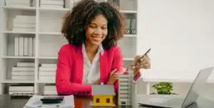 Pessoa em ambiente de escritório, segurando uma caneta e modelos de prédios sobre a mesa ao lado de um notebook, em imagem que ilustra artigo sobre Conta PJ da Cora para imobiliárias, com boletos, cobranças automatizadas e fluxo de caixa integrados.