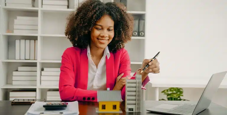 Pessoa em ambiente de escritório, segurando uma caneta e modelos de prédios sobre a mesa ao lado de um notebook, em imagem que ilustra artigo sobre Conta PJ da Cora para imobiliárias, com boletos, cobranças automatizadas e fluxo de caixa integrados.