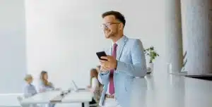 Pessoa em ambiente interno, segurando um smartphone em pé próximo a mesas de trabalho com outras pessoas ao fundo, em imagem que ilustra artigo sobre gestão moderna para imobiliárias: automações, cobranças recorrentes e fluxo de caixa.