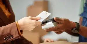Mãos de duas pessoas, uma segura o cartão para pagamento e a outra segura a maquininha, em imagem que ilustra artigo sobre como receber por cartão na empresa por diferentes meios de pagamento.