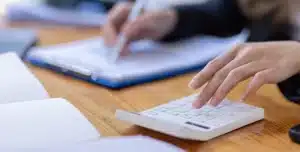Mão pressiona calculadora branca sobre mesa de madeira enquanto outra mão escreve em caderno ao fundo